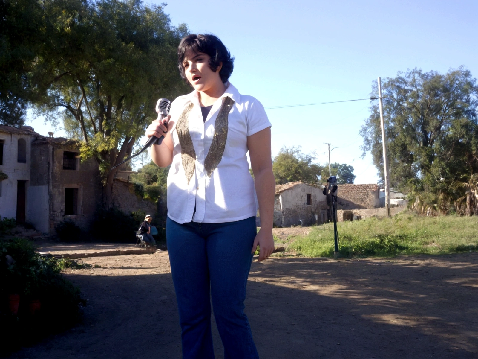 mujer cantando, vestida de chaqueta de jeans, remera blanca larga hasta los muslos, Oxford-style jeans from the 70s, cabello ondulado, recojido, con audifinos de vimvha, holding an old microphone from the 1940s, fondo de pradera, con pastos verdes, florores,