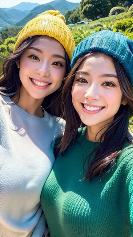 "Two women taking a selfie with a mountainous landscape in the background. One is wearing a blue beanie and the other is wearing a yellow beanie. Both are smiling brightly and wearing long-sleeve sweaters. Sunlight illuminates their faces, and the background features lush green trees and distant mountains under a clear sky, creating a beautiful scenic view."