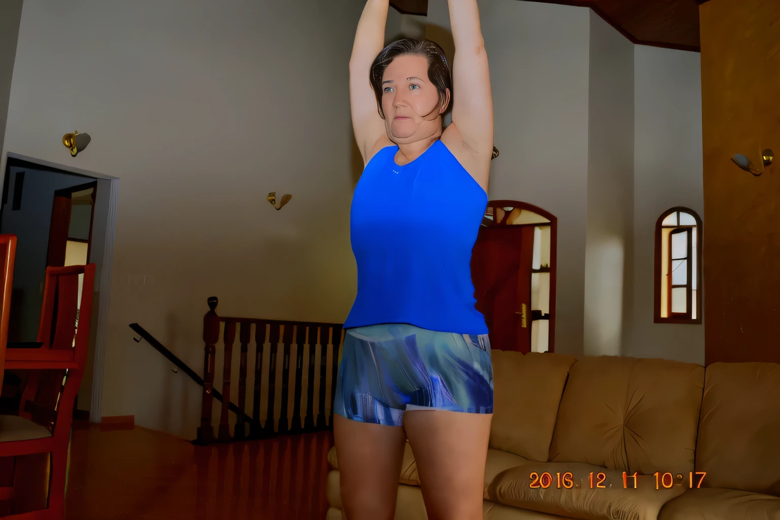 woman in blue top and shorts holding up a wii controller, sport bra and dark blue shorts, hunched shoulders, back arched, shoulders can be seen, waist reaching ponytail, upper body shot, indoor picture, in good physical shape, posing and flexing, fit pic, arms to side, wearing fitness gear, standing athletic pose, working out, sport bra and shorts