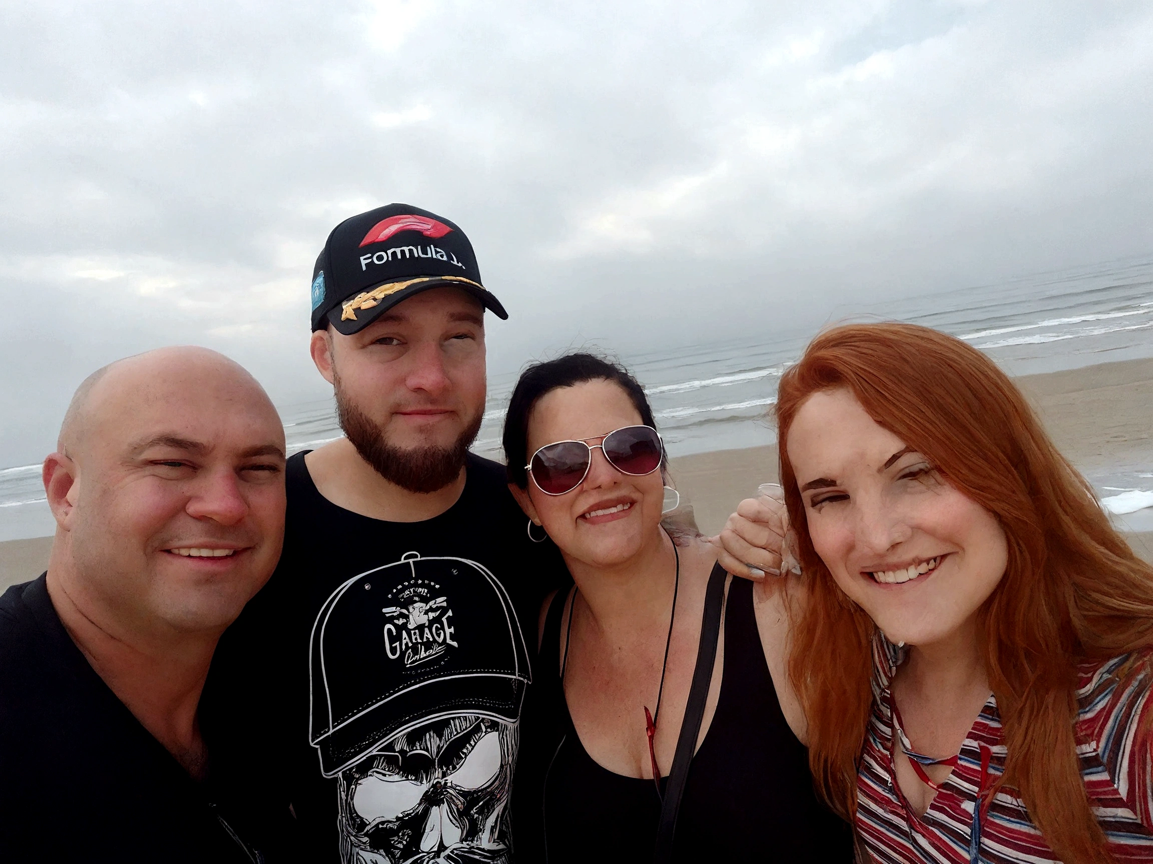 There are four people together on the beach, a man in a cap, a bald man, a woman with black hair and sunglasses and a young woman with red hair. On the beach, vacation photo, beach photo, standing near the beach, in the sea, on a beach, on a beach, on a sunny beach, on a beach, on the beach, on the beach, on the ocean, family photo, happy family