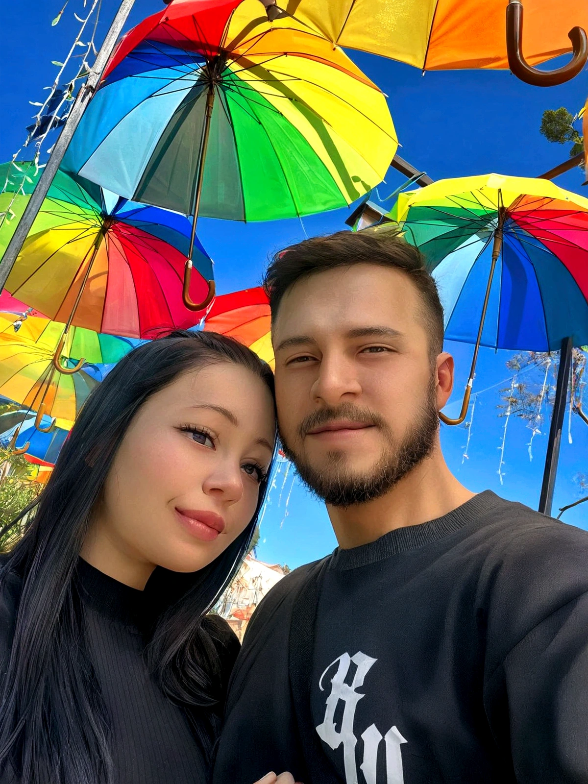 A young couple share a warm smile, enjoying the outdoor atmosphere on what appears to be a sunny day. The man has a full beard and is dressed casually in a black t-shirt, while the woman sports long black hair and a matching dark blouse. Their joyful interaction suggests a moment of shared happiness in a colorful and loving environment in the Disney Pixar style, high quality, better quality.
