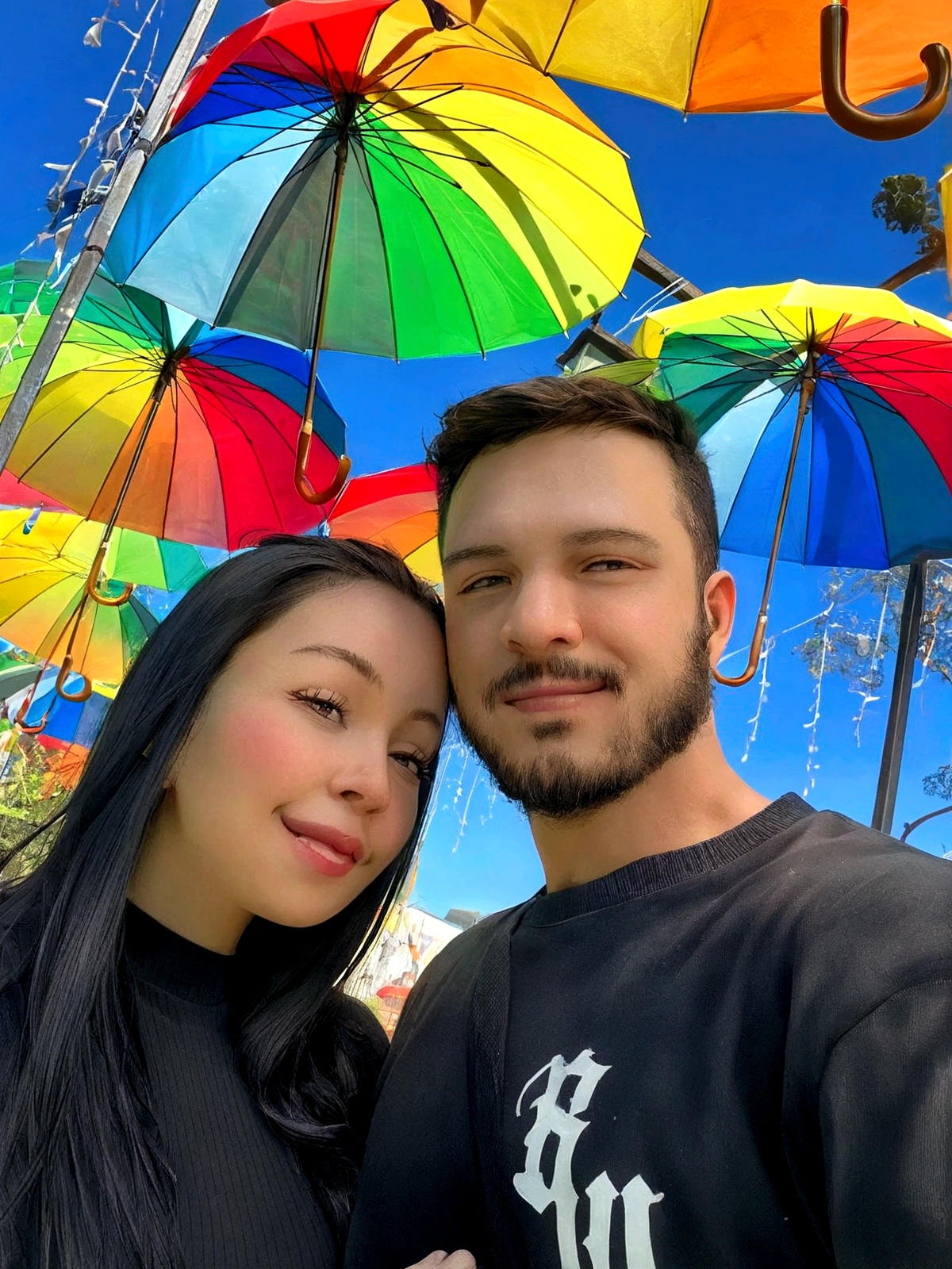 A young couple share a warm smile, enjoying the outdoor atmosphere on what appears to be a sunny day. The man has a full beard and is dressed casually in a black t-shirt, while the woman sports long black hair and a matching dark blouse. Their joyful interaction suggests a moment of shared happiness in a colorful and loving environment in the Disney Pixar style, high quality, better quality.
