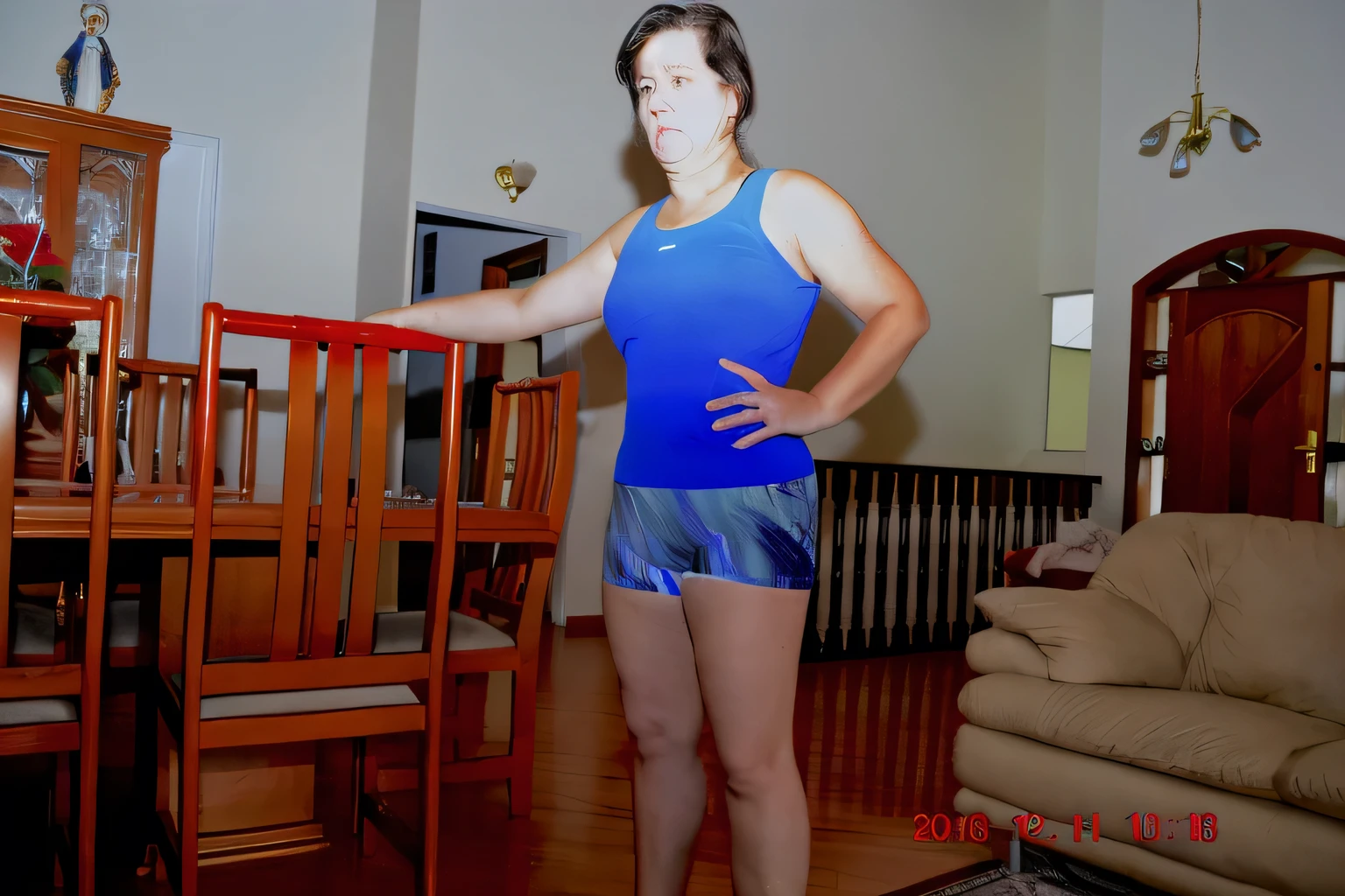 there is a woman standing in a living room with a wooden table, fit pic, in good physical shape, wearing track and field suit, wearing fitness gear, sport bra and dark blue shorts, short robust woman, standing athletic pose, wearing leotard, full body wide shot, indoor picture, distant full body shot, side pose, distant full body view