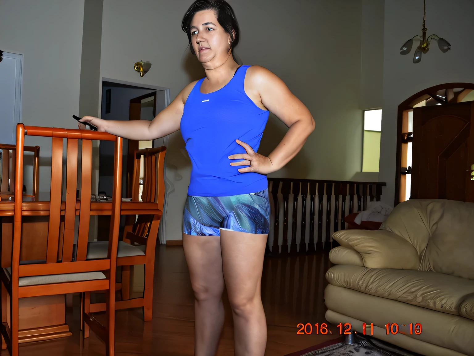 there is a woman standing in a living room with a chair, sport bra and dark blue shorts, short robust woman, she is about 45 years old, 40 years woman, sport bra and shorts, she is about 45 years old, in shorts, fit pic, 45 year brazilian mother, she is about 45 years old, photo from the back