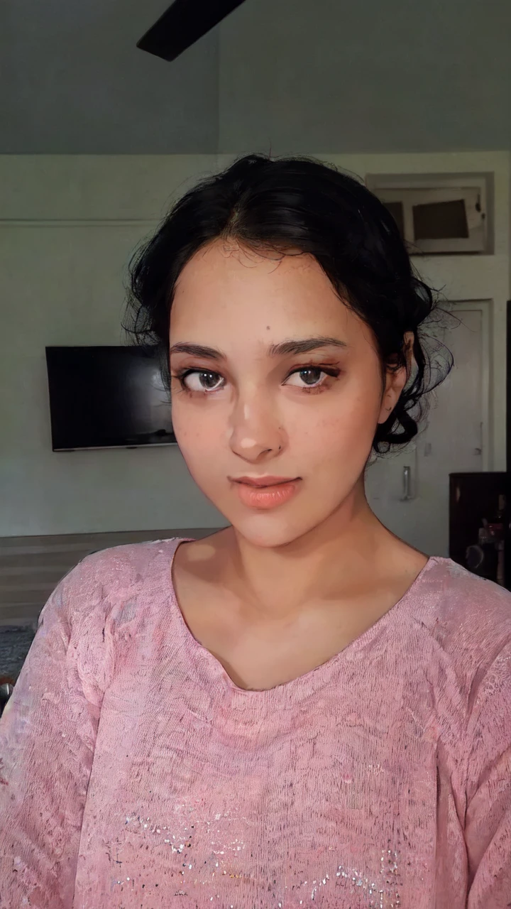 A young woman with a pink shirt and black hair is standing in front of a monitor or TV screen. The woman appears to be in her early twenties and has a feminine appearance. The photo also includes a picture or frame hanging on the wall in the background. The woman''s face is the main focus of the image, with her pink shirt standing out against the dark background. She has a neutral expression, with her eyes and mouth clearly visible. The overall color scheme of the image includes shades of pink, black, and brown.
