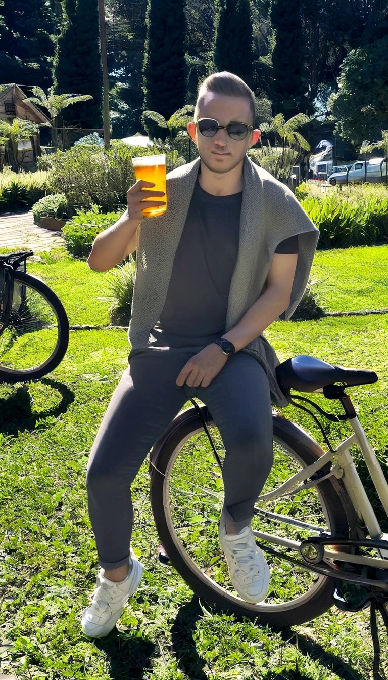 Slim man sitting on a bicycle with a glass of beer in his right hand. blonde hair and white beauty. Black t-shirt, gray pants and white sneakers.