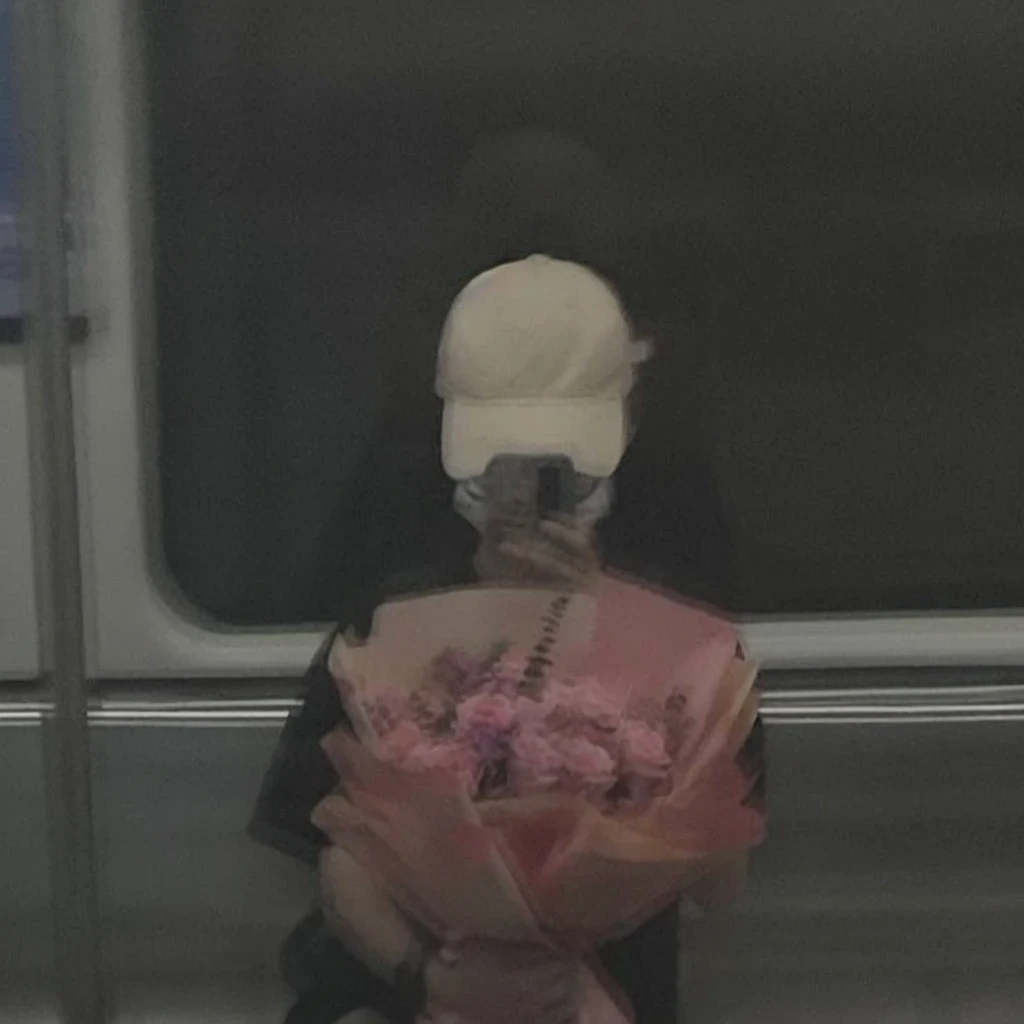 Prompt:
"Reflection of a person on a subway window, holding a bouquet of pink flowers. The person is wearing a white cap and a black outfit, partially obscured by the reflection and dim lighting. The background shows a blurred, dark subway interior, giving a moody and introspective ambiance. The bouquet is wrapped in pink paper, adding a soft, delicate contrast to the otherwise dark and muted tones of the scene."
Detailed Breakdown:
Setting: Subway window with reflection, dark interior.
Subject: Person holding a bouquet, wearing a white cap and black outfit.
Lighting: Dim, moody, emphasizing the reflective surface.
Colors: Dark tones with a pop of pink from the flowers.
Mood: Introspective, calm, and somewhat melancholic.
Use these details to guide the stable diffusion model in creating an image with a similar vibe and composition.