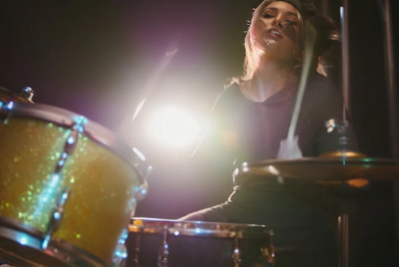 A young woman with vibrant silver hair, styled in a slightly messy pixie cut with the top slightly longer and the sides and back undercut, wearing impressive eyeliner, with a calm and neutral expression, striking green eyes, and light freckles scattered across her face. She is playing drums with a warm lighting that gives her skin a slightly glossy appearance, set against an abstract background with soft, warm tones and a few silver splatters to match her hair. She is wearing a black, rock fashion outfit with subtle silver accents. The overall mood is modern, slightly ethereal, and eye-catching.