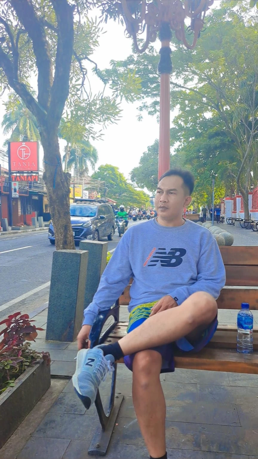 realistic caricature of a 35 year old Indonesian Muslim man wearing glasses sitting on a high wooden bench holding a cup of hot coffee. Beside him is a table containing a plate of fried tofu on a black background, taken in a photo studio