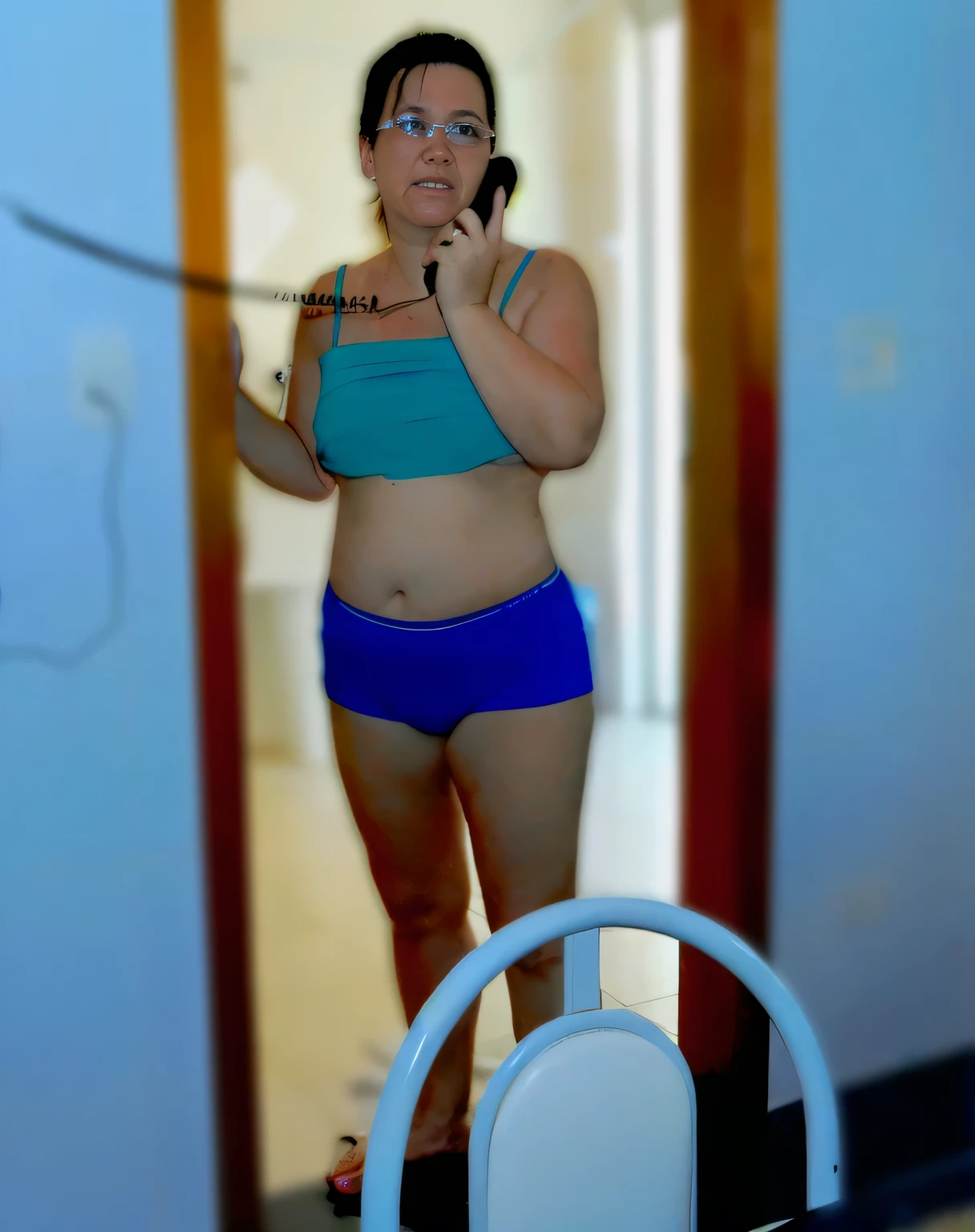 there is a woman in a blue bikini talking on a cell phone, fit pic, sport bra and dark blue shorts, mirror selfie, standing in front of a mirror, in bathroom, leaked photo, middle shot waist up, distorted photo, in good physical shape, wearing a camisole and shorts, waist - shot, sport bra and shorts, slim