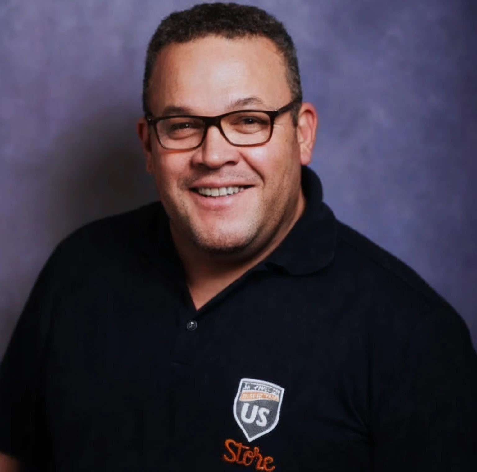 A dark man with curly hair, black t-shirt with writing "US" in orange, square and smiling glasses