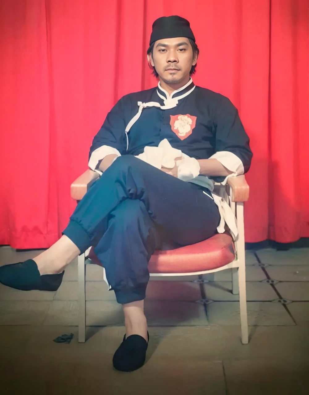 Realistic 3D caricature image of a full body Indonesian man sitting on a chair with a red curtain background. He is wearing traditional Chinese clothing, long-sleeved black cheongsam (sleeves rolled up to the elbows), shirt edges with white stripes on the edges and sleeves, shirt collar edges with stripes white. He wore a black skullcap. His hands were folded in his lap. He wore plain black joggers, white socks, and black traditional Chinese slip-on shoes (Wing Chun shoes) that matched his outfit. The man wore a calm and serious expression. The chair he sat on was red, possibly made of cloth or foam, with red armrests. He sat with his legs crossed, left leg over right. The backdrop features bright red curtains that stretch from top to floor. The floor visible below has a tile pattern, perhaps ceramic or tile, with some visible stains or dirt. Detailed HD quality cinematic 3D rendering.