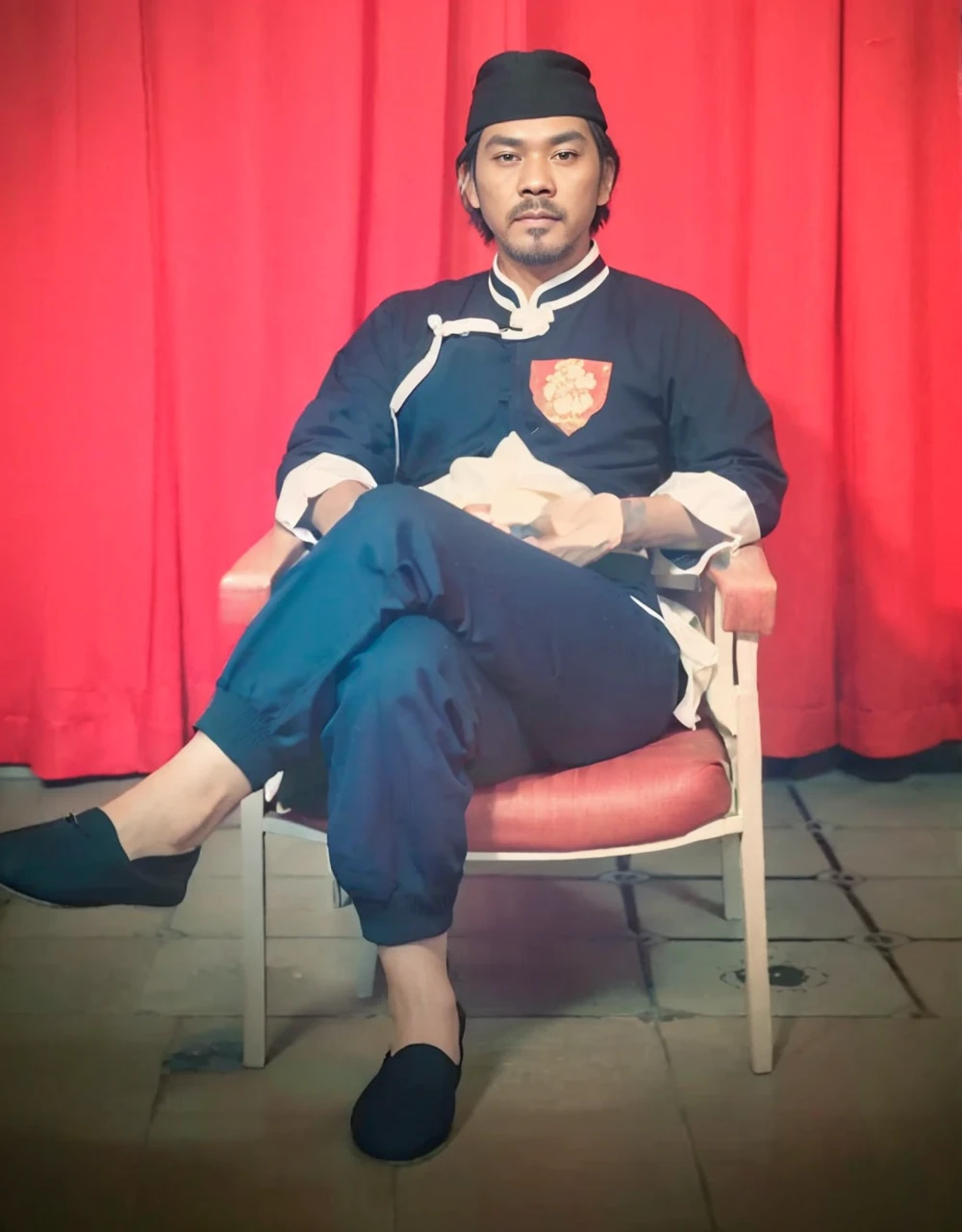 Realistic 3D caricature image of a full body Indonesian man sitting on a chair with a red curtain background. He is wearing traditional Chinese clothing, long-sleeved black cheongsam (sleeves rolled up to the elbows), shirt edges with white stripes on the edges and sleeves, shirt collar edges with stripes white. He wore a black skullcap. His hands were folded in his lap. He wore plain black joggers, white socks, and black traditional Chinese slip-on shoes (Wing Chun shoes) that matched his outfit. The man wore a calm and serious expression. The chair he sat on was red, possibly made of cloth or foam, with red armrests. He sat with his legs crossed, left leg over right. The backdrop features bright red curtains that stretch from top to floor. The floor visible below has a tile pattern, perhaps ceramic or tile, with some visible stains or dirt. Detailed HD quality cinematic 3D rendering.