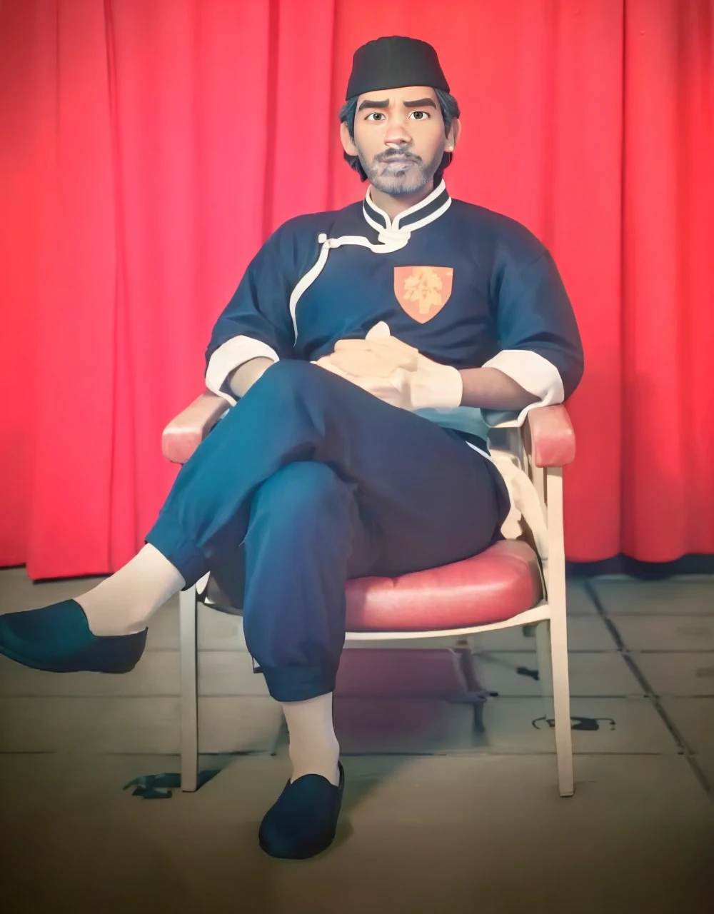 Realistic 3D caricature image of a full body Indonesian man sitting on a chair with a red curtain background. He is wearing traditional Chinese clothing, long-sleeved black cheongsam (sleeves rolled up to the elbows), shirt edges with white stripes on the edges and sleeves, shirt collar edges with stripes white. He wore a black skullcap. His hands were folded in his lap. He wore plain black joggers, white socks, and black traditional Chinese slip-on shoes (Wing Chun shoes) that matched his outfit. The man wore a calm and serious expression. The chair he sat on was red, possibly made of cloth or foam, with red armrests. He sat with his legs crossed, left leg over right. The backdrop features bright red curtains that stretch from top to floor. The floor visible below has a tile pattern, perhaps ceramic or tile, with some visible stains or dirt. Detailed HD quality cinematic 3D rendering.