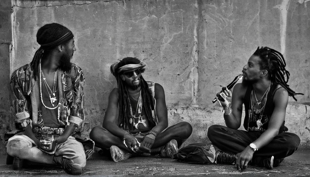 Jamaican Reggae band members are relaxing outside of a concert hall, having a break time near a parking lot. Drinking and smoking. Rasta style. Dreadlocks. Bandana. Black and white with natural lighting.