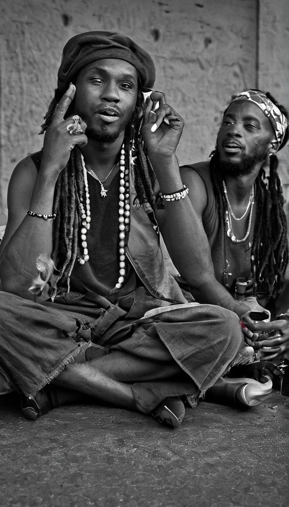 Three Jamaican Reggae band members are relaxing outside of a concert hall, having a break time near a parking lot. Drinking and smoking. Rasta style. Dreadlocks. Bandana. Black and white with natural lighting.