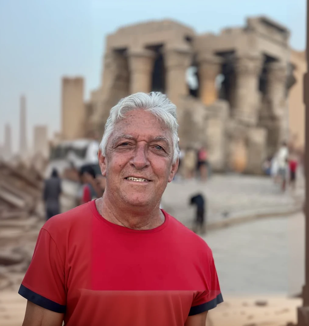 there is a man with red t-shirt that is standing, he is about 70 years old, ruins in the background, egypt