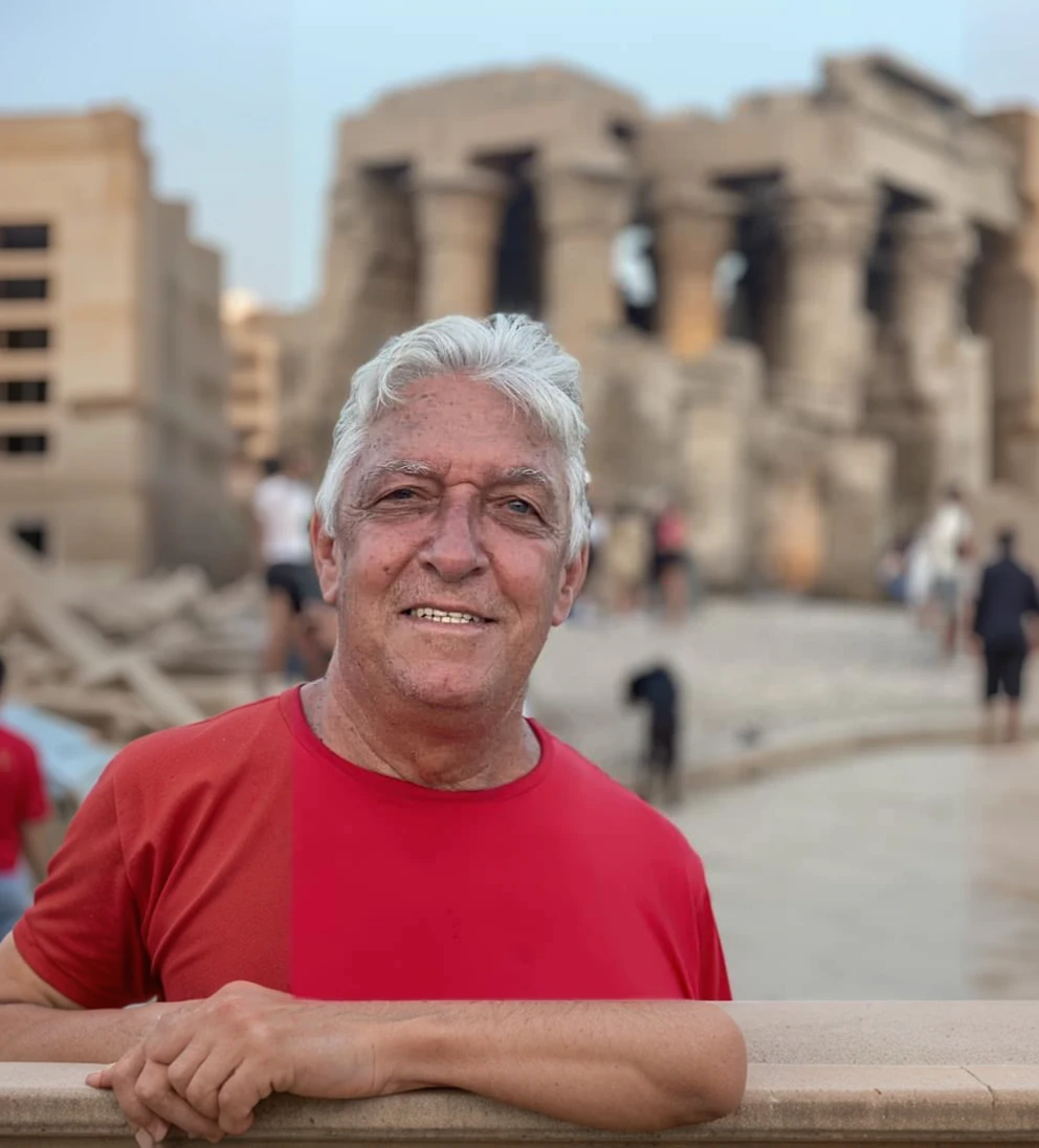 there is a man with red t-shirt that is standing, he is about 70 years old, ruins in the background, egypt
