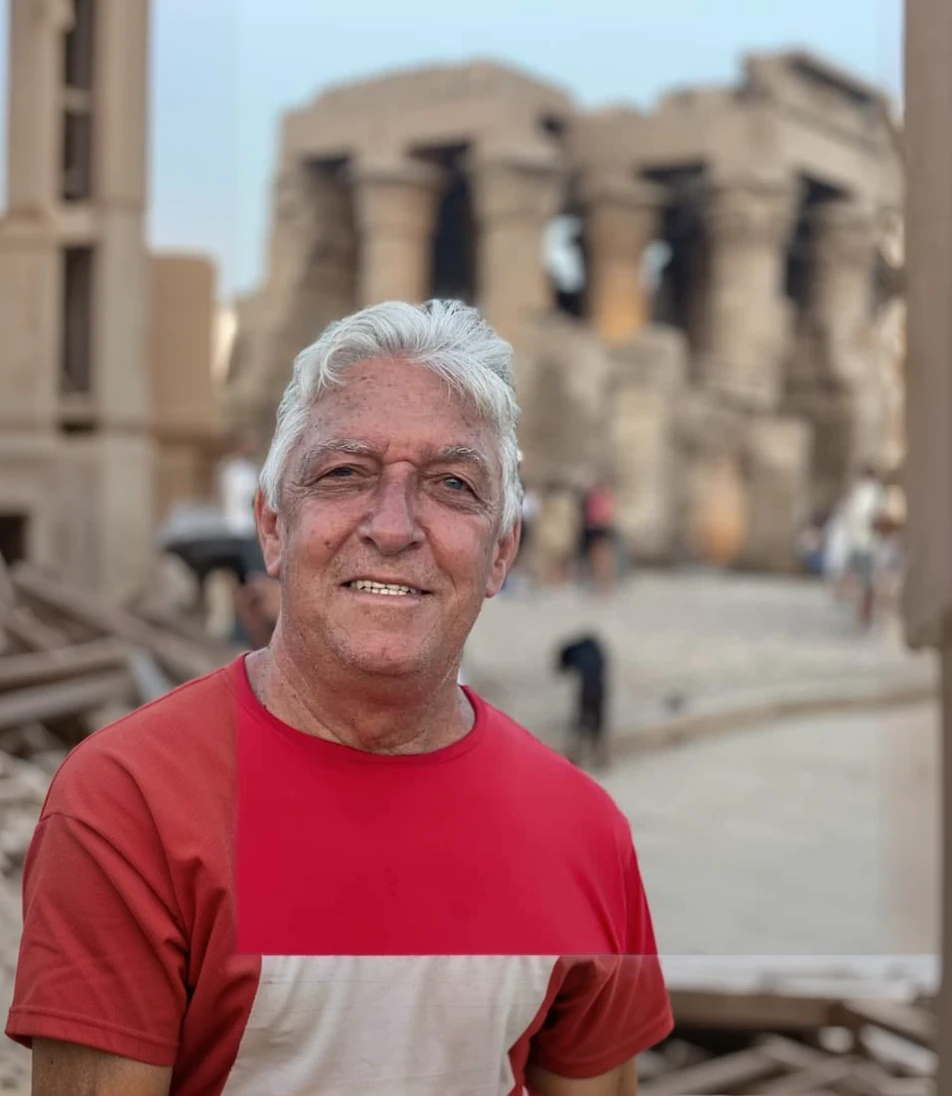 an old man with red t-shirt that is standing, he is about 70 years old, ruins in the background, egypt