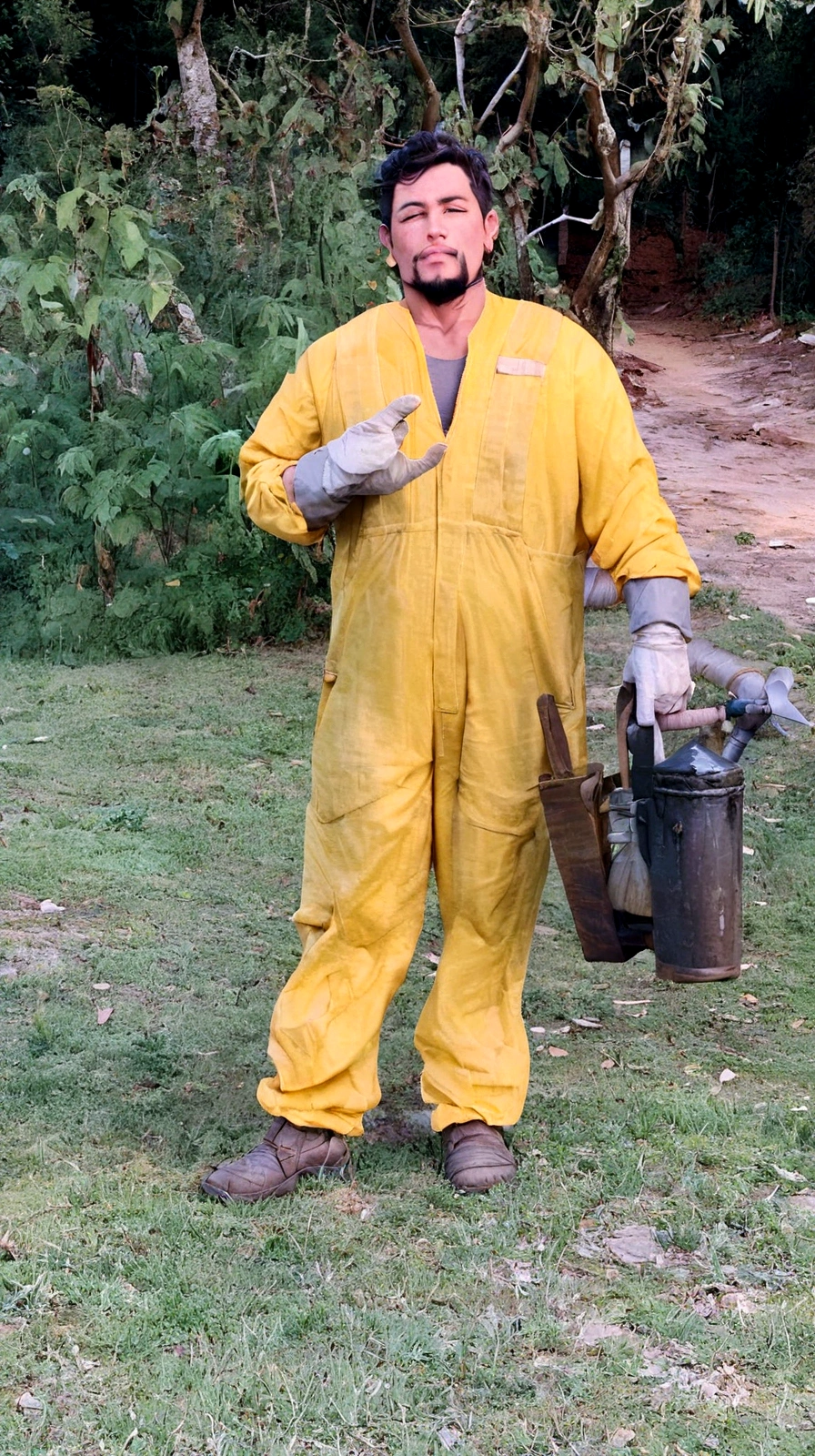 naughty man, Not too thin, black hair and goatee, dressed in yellow beekeeper overalls, drilling a beekeeping fumigator, making a horn sign with the right hand.