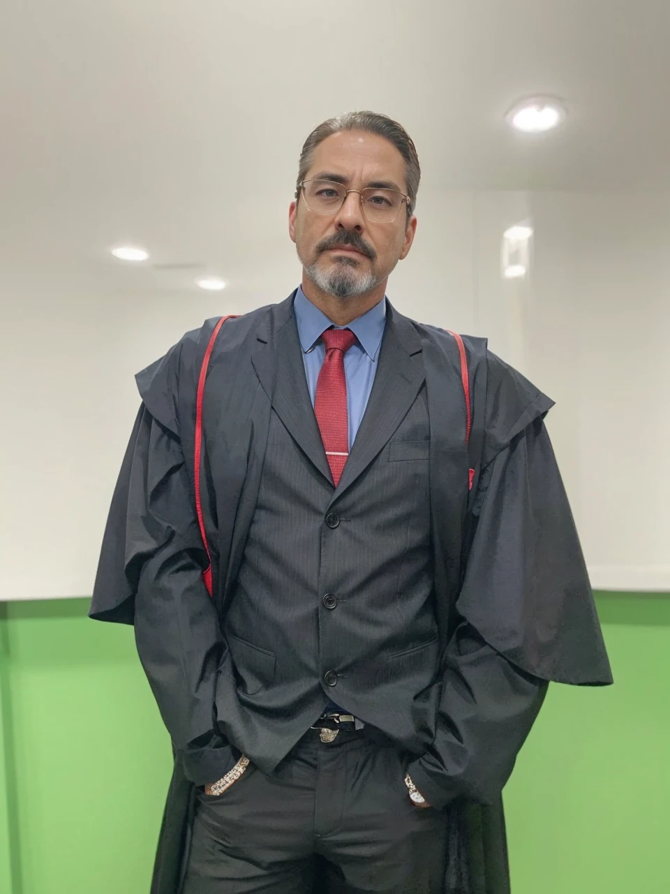 into the image, a man is dressed impeccably as a prosecutor. He wears a black toga with red details on the shoulders., indicating your position of authority in the judicial system. Your face has an oval shape and a well-defined bone structure., conveying seriousness and authority. Her skin tone is swarthy, contrasting elegantly with his gray beard and hair. The hair is short, gray and carefully combed back, giving a sophisticated and professional look. Your beard is well trimmed, outlining the chin line, and is inspired by the style of the character Tony Stark, not connecting to the mustache. The mustache is thin, well outlined and separated from the beard. Your eyebrows are black, thick and slightly arched, standing out against dark skin tone and providing intensity to your look. He wears glasses with a rectangular dark metal frame., discreet but functional, that frame your face without distracting from your serious appearance. Underneath the toga, he is dressed in a black suit, a light blue shirt and a red tie, adding a touch of sophistication and formality. Your posture, with hands in pockets, suggests a combination of confidence and relaxation, balancing the seriousness of the occasion with a relaxed attitude. The background of the image is simple, with a white wall and a green stripe at the bottom, keeping the focus entirely on the prosecutor. The man's appearance clearly conveys the image of a serious and respectable professional, ready to perform their duties with dignity and competence.