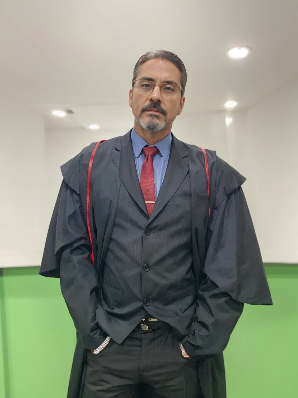 into the image, a man is dressed impeccably as a prosecutor. He wears a black toga with red details on the shoulders., indicating your position of authority in the judicial system. Your face has an oval shape and a well-defined bone structure., conveying seriousness and authority. Her skin tone is swarthy, contrasting elegantly with his gray beard and hair. The hair is short, gray and carefully combed back, giving a sophisticated and professional look. Your beard is well trimmed, outlining the chin line, and is inspired by the style of the character Tony Stark, not connecting to the mustache. The mustache is thin, well outlined and separated from the beard. Your eyebrows are black, thick and slightly arched, standing out against dark skin tone and providing intensity to your look. He wears glasses with a rectangular dark metal frame., discreet but functional, that frame your face without distracting from your serious appearance. Underneath the toga, he is dressed in a black suit, a light blue shirt and a red tie, adding a touch of sophistication and formality. Your posture, with hands in pockets, suggests a combination of confidence and relaxation, balancing the seriousness of the occasion with a relaxed attitude. The background of the image is simple, with a white wall and a green stripe at the bottom, keeping the focus entirely on the prosecutor. The man&#39;s appearance clearly conveys the image of a serious and respectable professional, ready to perform their duties with dignity and competence.