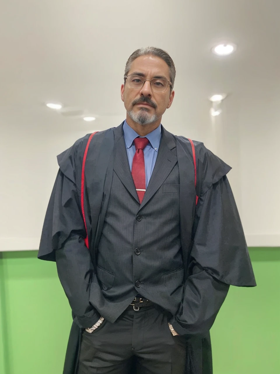 into the image, a man is dressed impeccably as a prosecutor. He wears a black toga with red details on the shoulders., indicating your position of authority in the judicial system. Your face has an oval shape and a well-defined bone structure., conveying seriousness and authority. Her skin tone is swarthy, contrasting elegantly with his gray beard and hair. The hair is short, gray and carefully combed back, giving a sophisticated and professional look. Your beard is well trimmed, outlining the chin line, and is inspired by the style of the character Tony Stark, not connecting to the mustache. The mustache is thin, well outlined and separated from the beard. Your eyebrows are black, thick and slightly arched, standing out against dark skin tone and providing intensity to your look. He wears glasses with a rectangular dark metal frame., discreet but functional, that frame your face without distracting from your serious appearance. Underneath the toga, he is dressed in a black suit, a light blue shirt and a red tie, adding a touch of sophistication and formality. Your posture, with hands in pockets, suggests a combination of confidence and relaxation, balancing the seriousness of the occasion with a relaxed attitude. The background of the image is simple, with a white wall and a green stripe at the bottom, keeping the focus entirely on the prosecutor. The man's appearance clearly conveys the image of a serious and respectable professional, ready to perform their duties with dignity and competence.