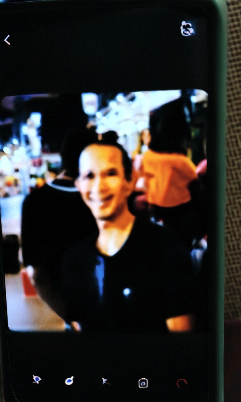 there is a cell phone with a photo of a man, very very low quality picture, heavy blurry jpeg artifact, blurry image, blurry face, he is wearing a black t-shirt, zoomed out shot, very very low quality, appearing in a mall, detailed blurred face, very blurry, blurry photo, person in foreground