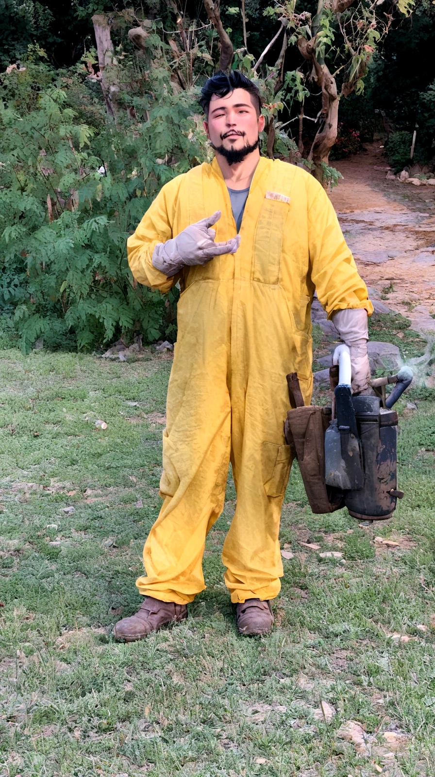man with light skin, black hair and goatee, in yellow beekeeper overalls holding a fumigator in his right hand
