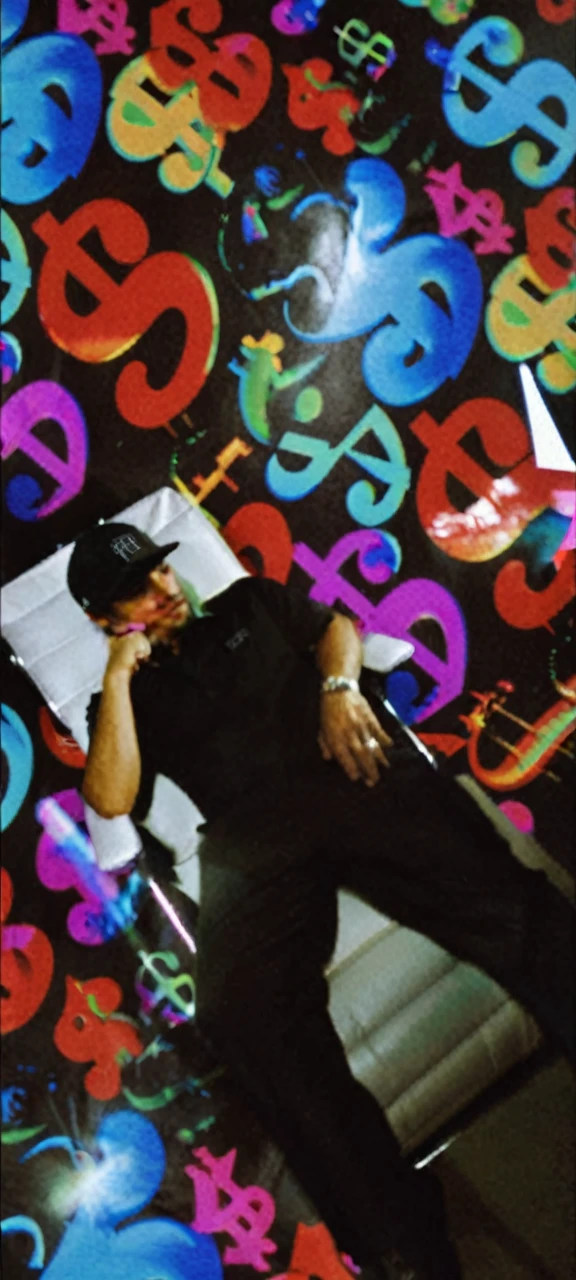 A handsome man wears a black cap with the monopoly logo, He wears black clothing and is sitting with a confident pose on a white leather executive chair with chrome details.