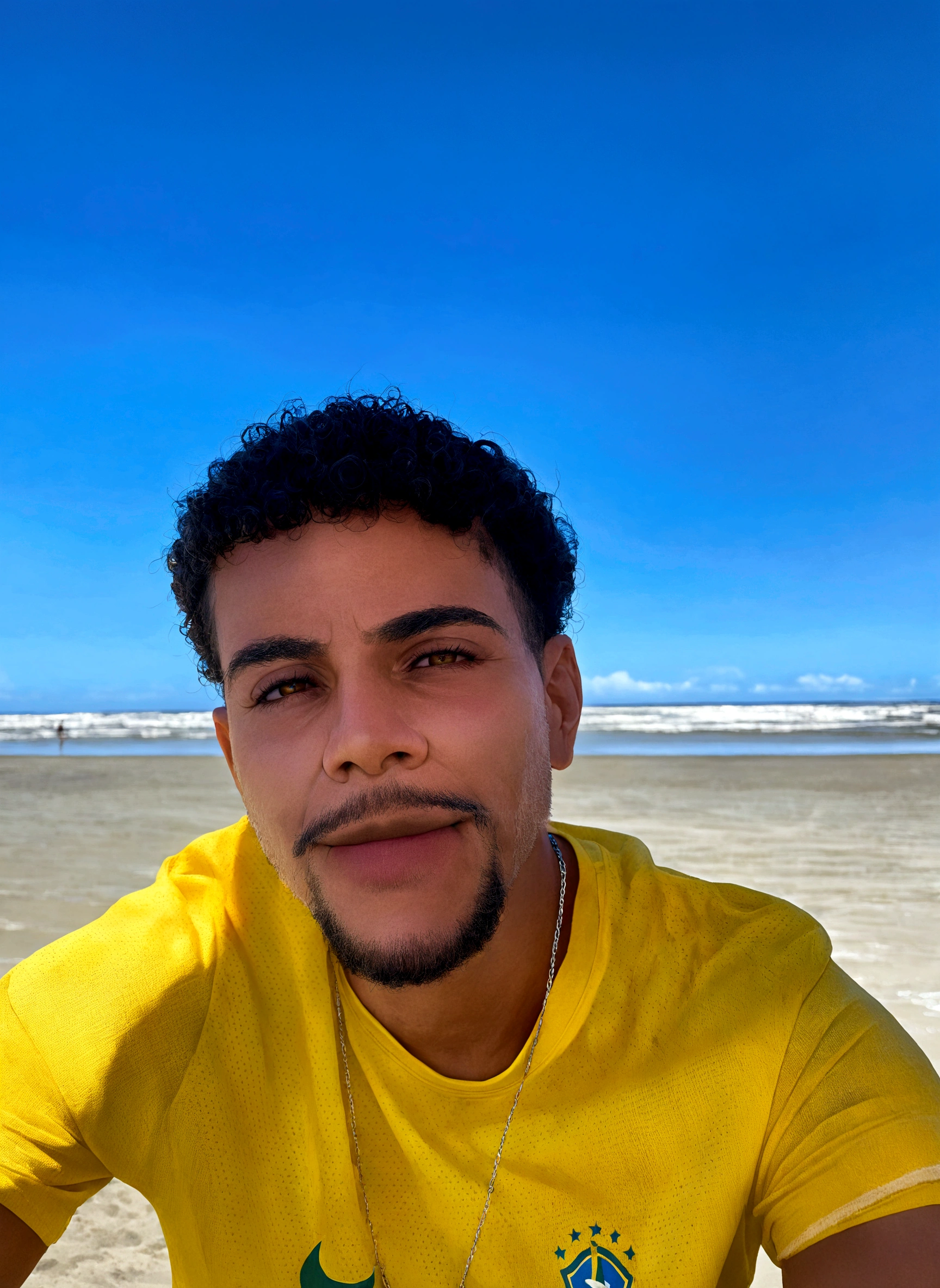 Man on the beach in a yellow t-shirt and chain, giving a smile, eyes browns, curly hair and mustache