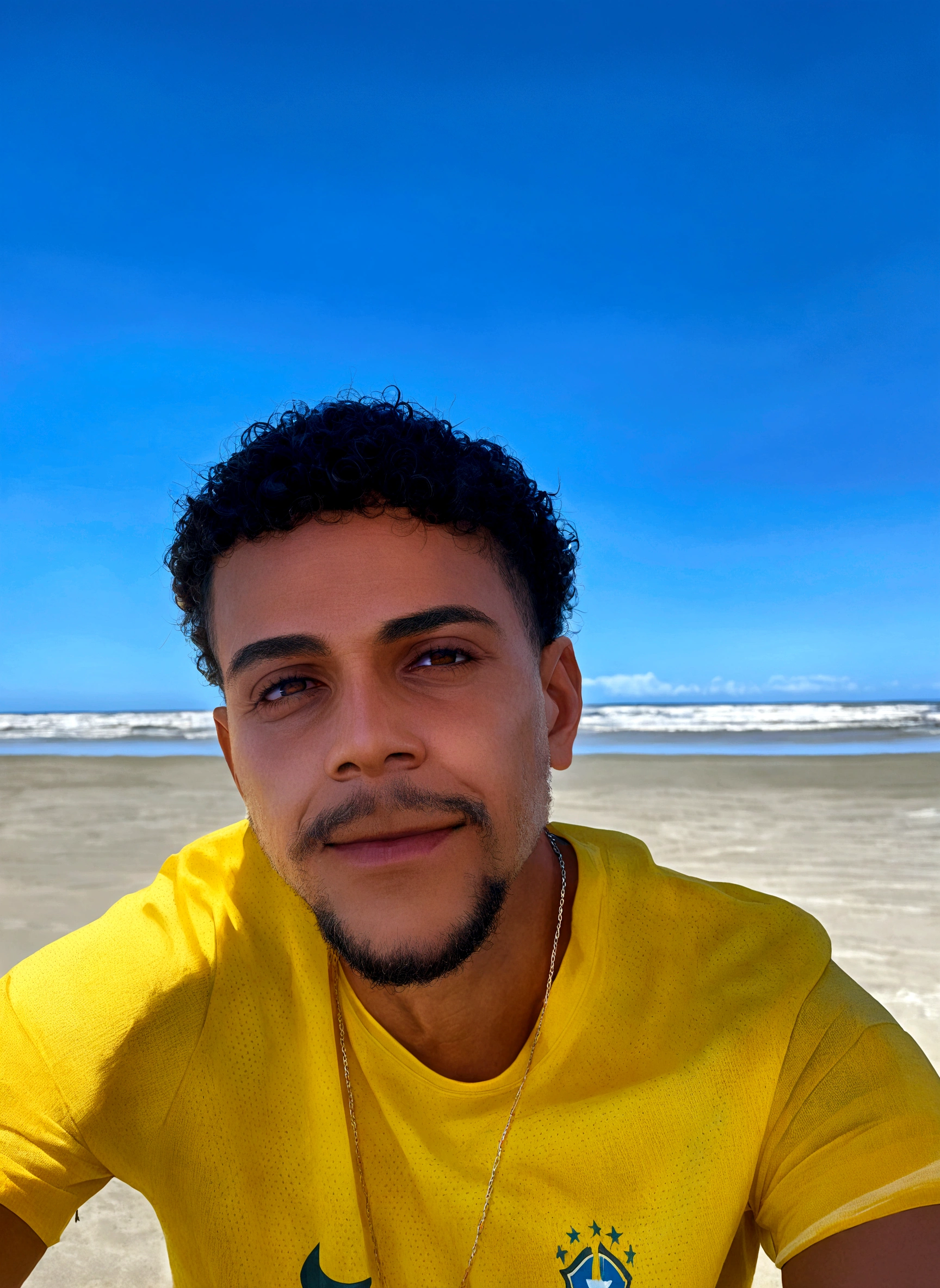 Man on the beach in a yellow t-shirt and chain, smiling, brown eyes, curly hair and mustache