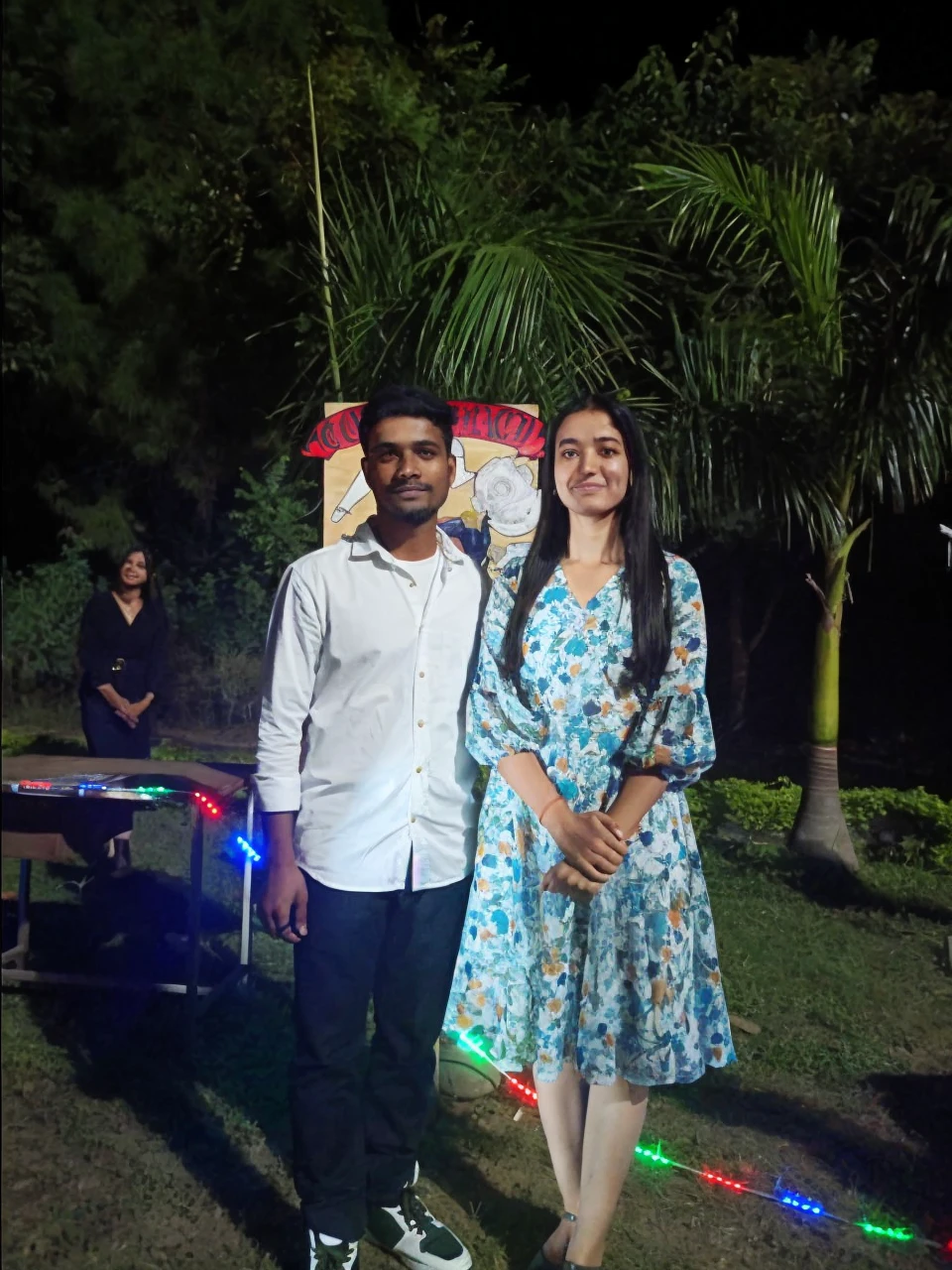 "A young man and woman standing side by side, facing the camera. The young man has short, neatly styled dark hair and a trimmed beard. He has a medium build and medium brown skin tone. He is wearing a white button-down shirt with the sleeves rolled up to his elbows and dark blue jeans. His shoes are white with green accents. He has a calm and confident expression, looking directly at the camera with dark, almond-shaped eyes, and his lips are neutral, giving a slight, relaxed smile.
The young woman has long, straight, dark hair that falls past her shoulders. She has an oval face with a smooth, light brown complexion. Her eyes are almond-shaped, dark brown, and expressive, framed by well-defined eyebrows. She has a gentle, warm smile with full lips. She is wearing a knee-length, floral print dress with shades of blue, white, and orange. The dress has long sleeves that are slightly puffed and a fitted waist. Her footwear consists of simple, flat shoes. She stands with her hands clasped in front of her, looking directly at the camera with a calm and serene expression.
Both individuals are standing close to each other, exuding a sense of comfort and familiarity. The lighting is soft and even, illuminating their faces clearly