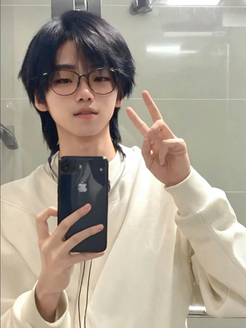Asian boy. Features: looking at the camera + Holding the cell phone with one hand and the other hand raised, with both fingers raised + thin face + delicate skin + delicate features + usa óculos