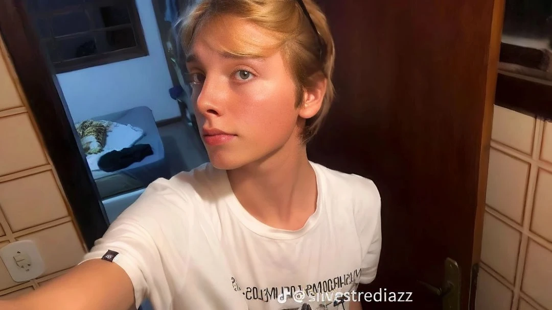 a blond boy taking a selfie with his cell phone in a room