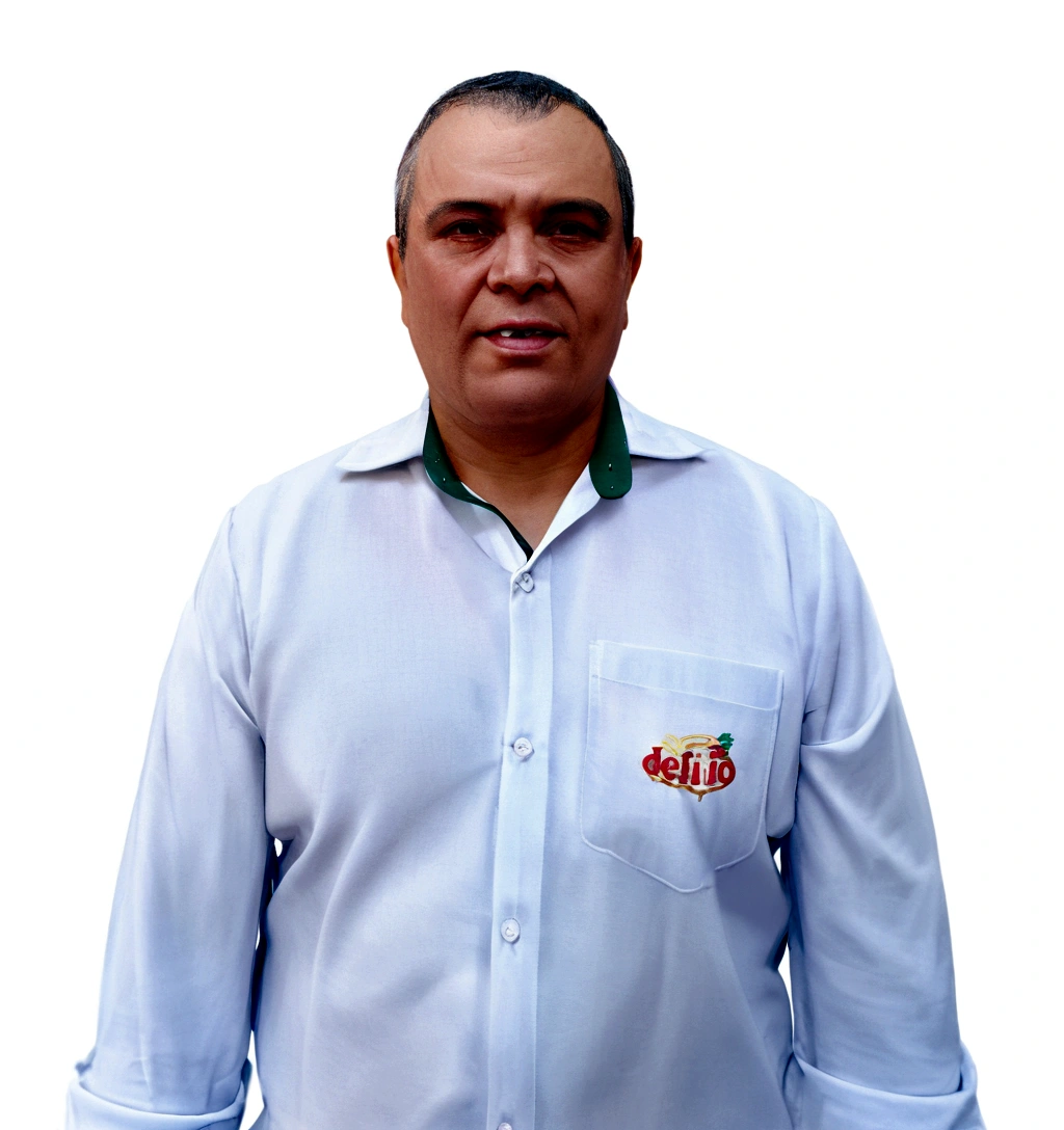 He is a man with fair skin, short dark hair, and some signs of baldness. He is wearing a white dress shirt with light buttons, open at the collar, revealing a green detail on the inside of the collar. On the shirt pocket, there is an embroidery with the "delrio" logo in red and yellow letters, accompanied by an image of a stylized fruit.