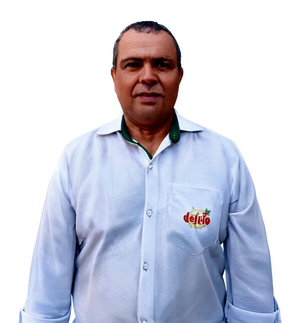 He is a man with fair skin, short dark hair, and some signs of baldness. He is wearing a white dress shirt with light buttons, open at the collar, revealing a green detail on the inside of the collar. On the shirt pocket, there is an embroidery with the "delrio" logo in red and yellow letters, accompanied by an image of a stylized fruit.