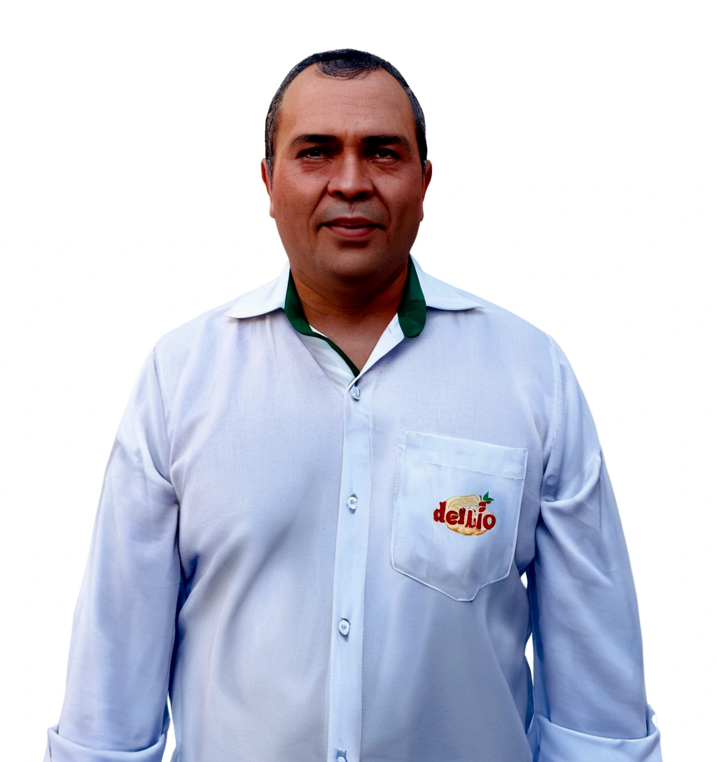 He is a man with fair skin, short dark hair, and some signs of baldness. He is wearing a white dress shirt with light buttons, open at the collar, revealing a green detail on the inside of the collar. On the shirt pocket, there is an embroidery with the "delrio" logo in red and yellow letters, accompanied by an image of a stylized fruit.