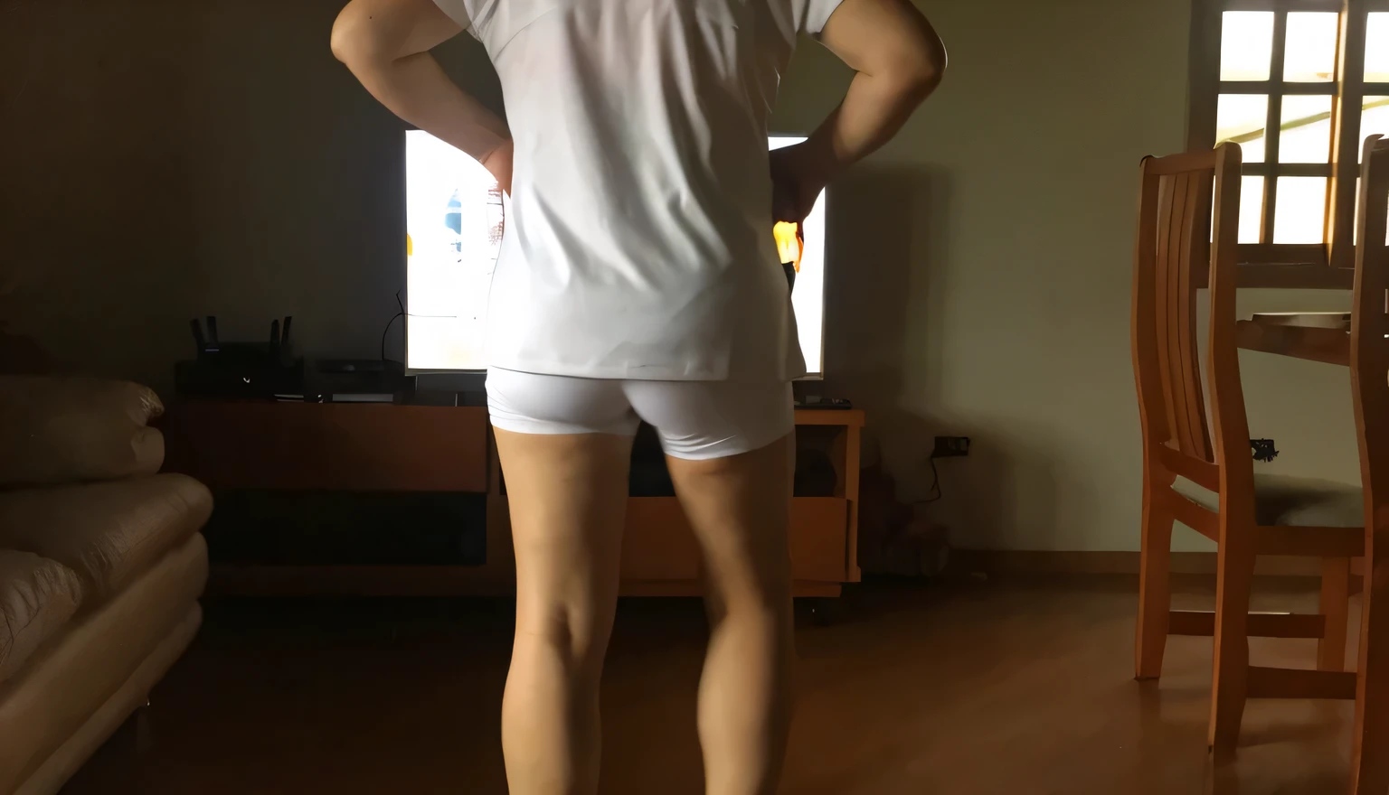 there is a man standing in front of a television in a living room, wearing white tights, legs visible, wearing shorts and t shirt, in shorts, long shot from the back, long shot from back, back view!!, centered full body rear-shot, shot from the back, rear-shot, distant full body view, sexy girl wearing shorts