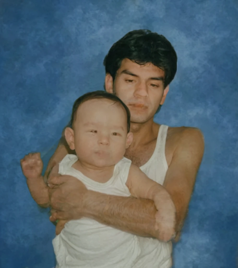 The image shows a man and a baby, both with dark hair, standing against a blue background. The man appears to be wearing a white shirt