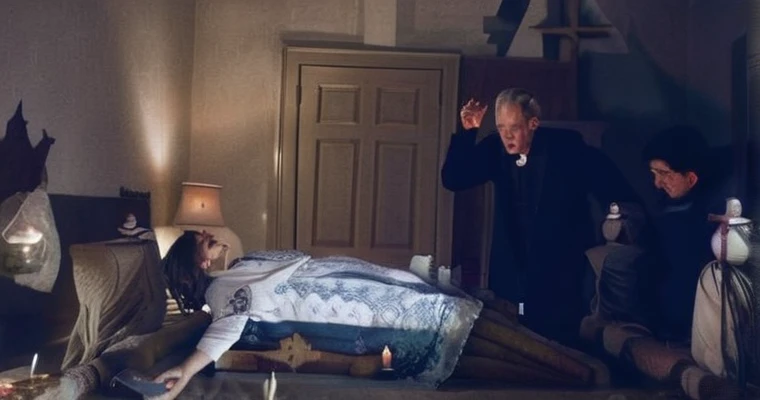 The girl Regan levitates above his bed, with a demonic expression, while priests recite prayers with raised crosses. The room is in chaos, with objects floating and lights flashing. The scene is intense and dramatic.