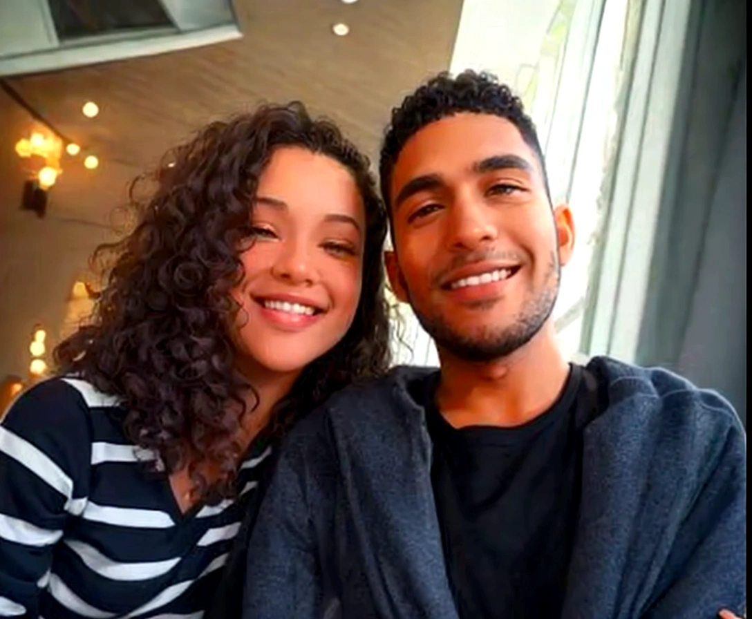 couple is smiling and happy The man is wearing a black t-shirt under a dark gray coat Hair: Short and curly, black color, brown skin. The woman is wearing a black and white striped blouse Hair: Curly and voluminous, brown color, white skin.
. Both are on a wooden walkway with flowers on the side. In the background there are white tables and chairs, above there are branches of a tree.