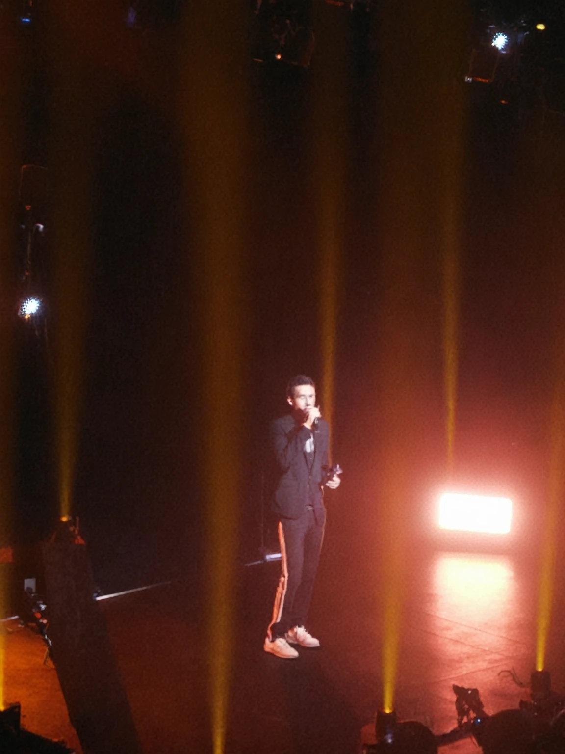 there is a man standing on stage with a microphone, on stage, on stage, tyler, Performance, very grainy, concert photo, concert, 3 AM, 3 AM, iPhone Capture, in Paris, Very very low quality image, photo [ Far ], paris 2010, Long-distance full-body photo, A bit vague, on stage, concert footage
