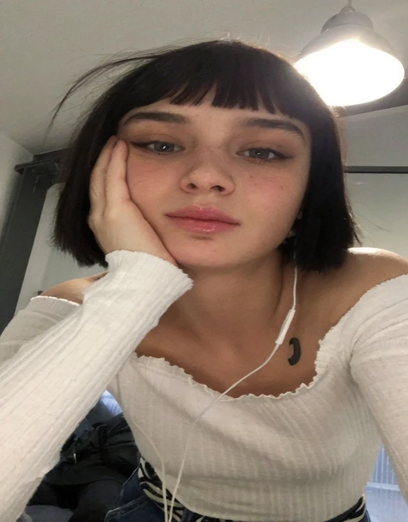 Pale teenage girl with black hair and bangs above her eyebrows, with small pink lips and a potato nose and light green eyes, taking photo with long t-shirt with one hand on left cheek