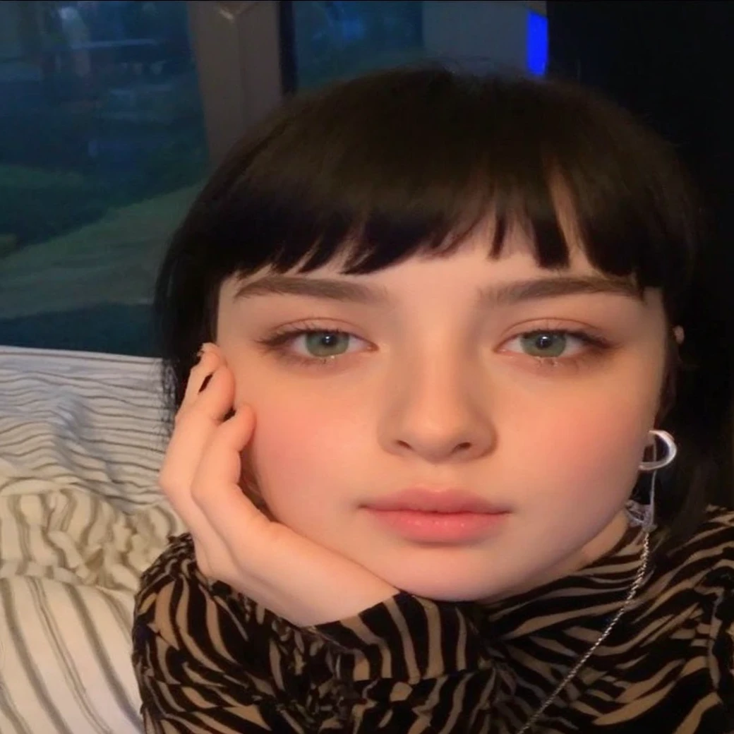 Teenage girl with clean pale skin and silky soft black hair, with small pink lips and a small, thin potato nose and blue-green eyes , taking photo looking at camera bored