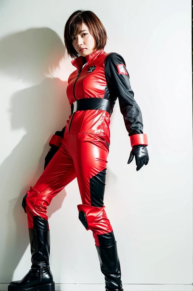 A woman in a red and black Sentai hero suit stands upright against a white wall. The suit is made of a shiny material, and she is wearing white gloves and yellow-red boots. The woman has short, shoulder-length hair and a natural look on her face.