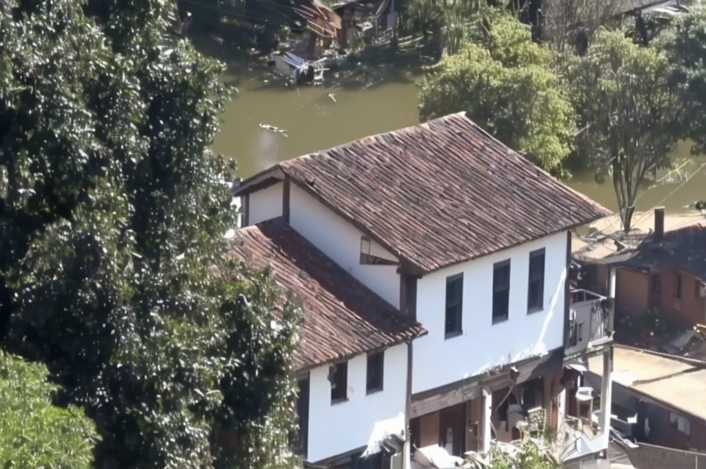 there is a white house with a brown roof and a brown roof, building along a river, colonial house in the background, small town house, small town, old house, stream flowing by the house, photo taken from above, gui guimaraes, viewed from the top, video still, close de cima, from above view, shot from 50 feet away