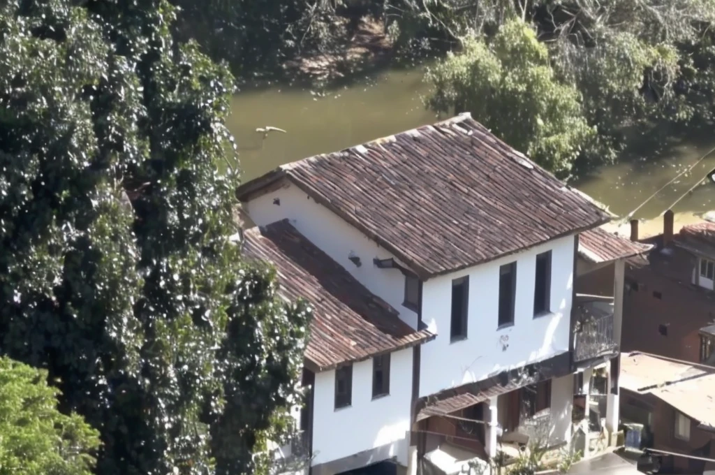 there is a white house with a brown roof and a brown roof, building along a river, colonial house in the background, small town house, small town, old house, stream flowing by the house, photo taken from above, gui guimaraes, viewed from the top, video still, close de cima, from above view, shot from 50 feet away