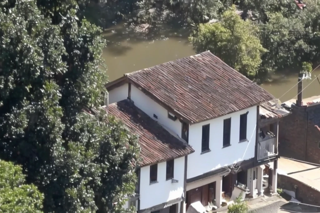 there is a white house with a brown roof and a brown roof, building along a river, colonial house in the background, small town house, small town, old house, stream flowing by the house, photo taken from above, gui guimaraes, viewed from the top, video still, close de cima, from above view, shot from 50 feet away