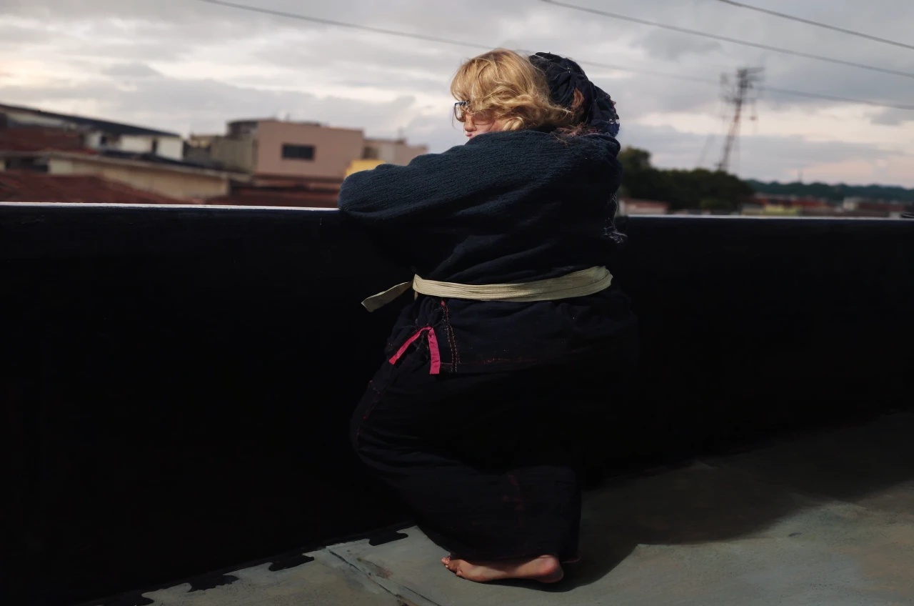 Slightly chubby blonde woman, of square glasses, blondie hair, clear green eyes, on the balcony looking at the sea to the horizon at sunset